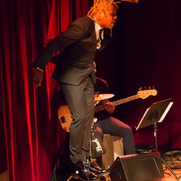 Omar Edwards appears to levitate vertically against stage right curtain wing in front of bass player. Omar, blonde braids, in black suit, white shirt black tie, black ankle high tap boots.