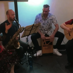 Flamenco Latino returns at Espresso 77 in Jackson Heights on 11/3/23. Image from 2022. Left to Right: Aurora Reyes, gray hair in bun at top of head, seated doing palmas, dressed in black costume with red polka dots, having ruffle with white polka dots and see-through black long sleeves, also wearing red kerchief with small white polka dots around neck, with ends going down center of chest; Danny Rivera, reddish hair, very receding hairline with sideburns, mustache and beard grown in about 3/8 inches, seated playing baritone saxophone, wearing dark blue shirt and black pants, Luis Osso, seated on top of cajón, playing face of cajón, short black hair about 1-1/2 inches on top of head, clipped short at sides with a little graying, wearing white short sleeve shirt with dark blue floral pattern, blue jeans, and white ankle high athletic shoes; Basilio Georges white, silver hair and goatee, playing flamenco guitar, wearing crimson t-shirt with his catch phrase “Still Can’t Git No Grindin’” in white text, with black vest, black pants, black athletic shoes.